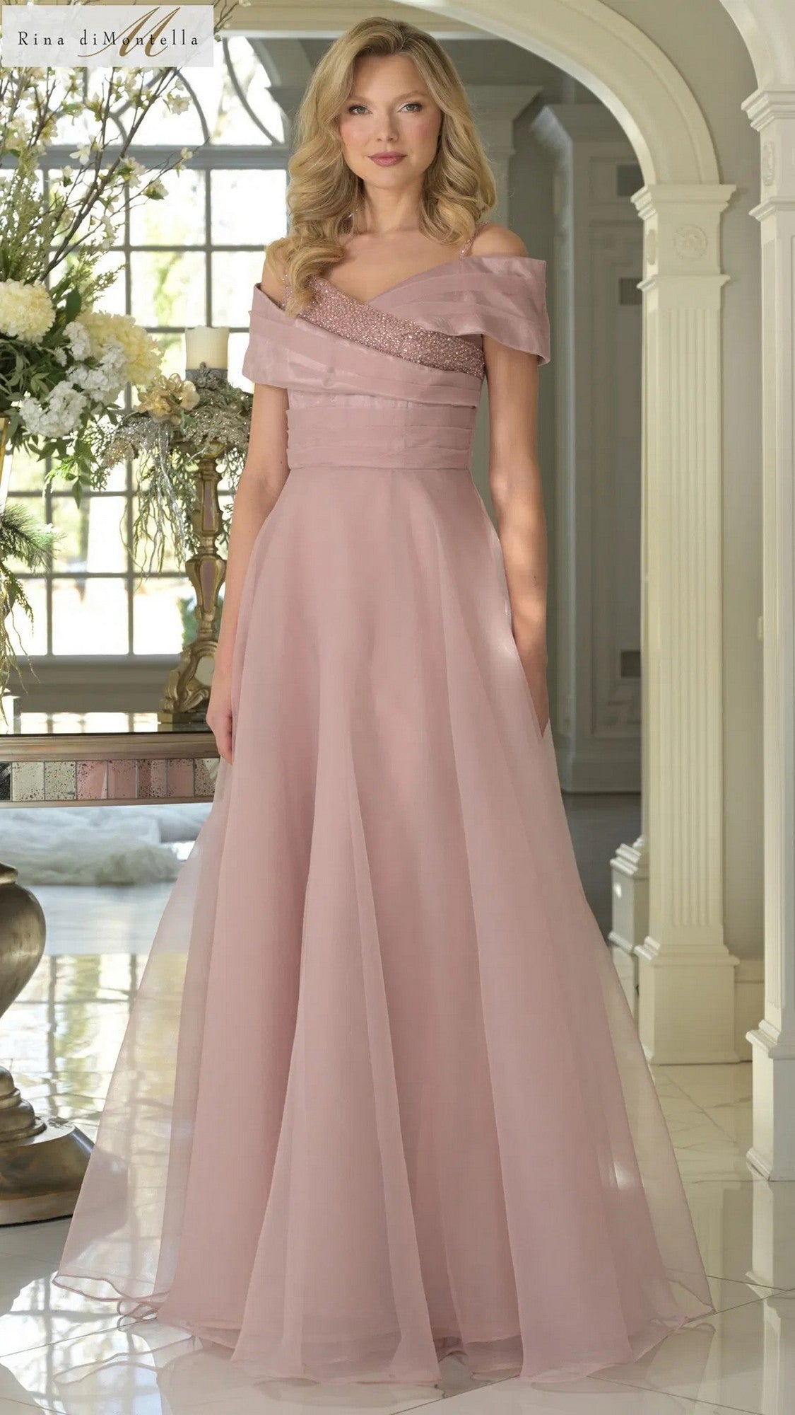 A woman stands indoors wearing the Long Formal Dress: Rina Di Montella RD3303 in Dusty Rose, a floor-length organza gown with an off-shoulder neckline and fitted bodice. Flowers and a window can be seen behind her.
